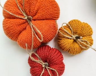 May include: Three knitted pumpkins in orange, yellow, and red with twine bows. The pumpkins are handmade and have a rustic, autumnal feel.