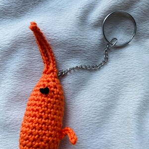 May include: A handmade orange crochet shrimp keychain with a black heart-shaped eye and small arms. The shrimp has a white and orange striped collar and is attached to a silver chain and keyring.