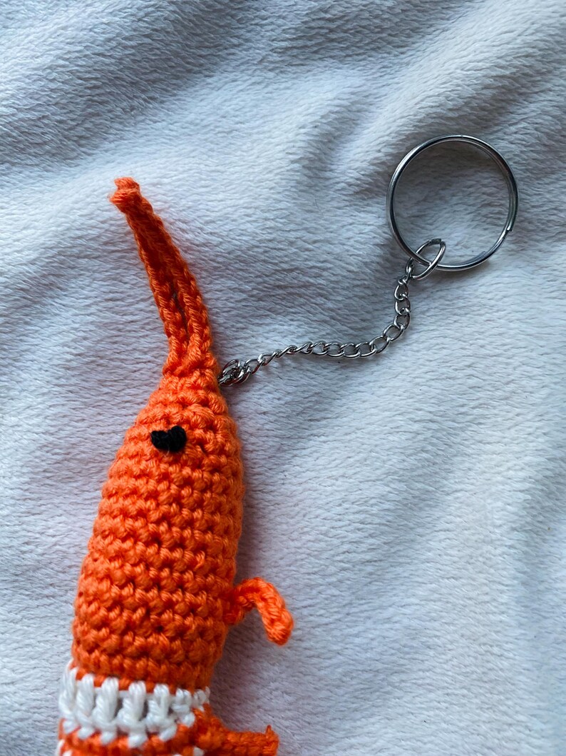 May include: A handmade orange crochet shrimp keychain with a black heart-shaped eye and small arms. The shrimp has a white and orange striped collar and is attached to a silver chain and keyring.