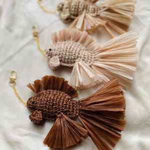 Può includere: Tre portachiavi a forma di pesce fatti a mano in varie tonalità di marrone e beige. Ogni pesce ha un corpo intrecciato, una coda sfrangiata e una catena color oro con chiusura. I portachiavi sono disposti su una superficie bianca e morbida.