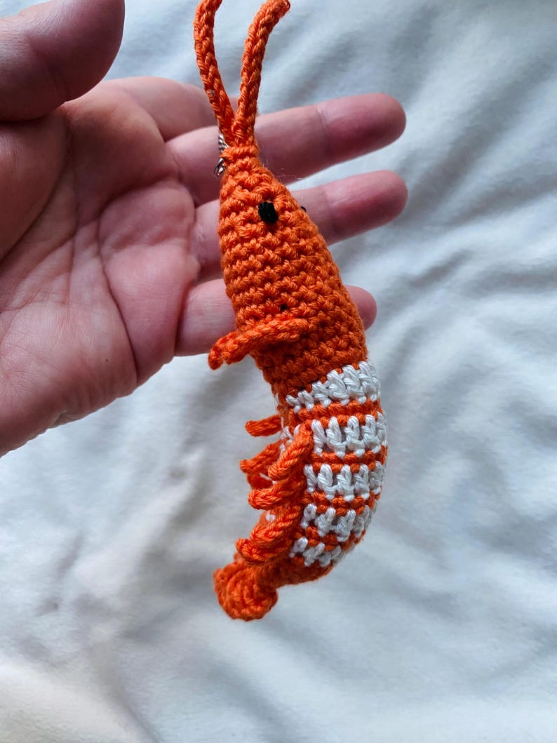 May include: A handmade orange crochet shrimp with white stripes on its tail. The shrimp has small black eyes and long orange antennae. This unique, whimsical item is perfect for ocean-themed decor or as a fun accessory.