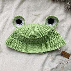 May include: A light green, crocheted frog hat with large, round eyes. The eyes are white with black centers and are attached to the top of the hat. The hat is handmade and has a small tag that says "Handmade with Love."