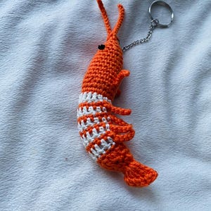 May include: A handmade orange crochet shrimp keychain with white stripes around the midsection. The shrimp has small black eyes, long antennae, and a silver chain attached to a keyring. The shrimp is made of yarn.