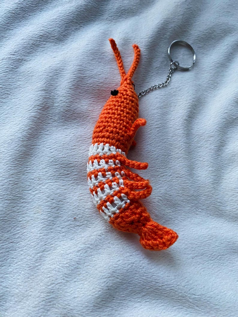 May include: A handmade orange crochet shrimp keychain with white stripes around the midsection. The shrimp has small black eyes, long antennae, and a silver chain attached to a keyring. The shrimp is made of yarn.