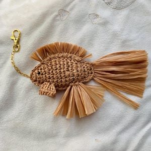 May include: A woven raffia fish keychain with a gold-colored clasp and chain. The fish has a textured body, a fringed tail, and fins. The keychain is a light brown color and is set against a white background.