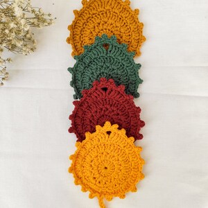 Puede incluir: Cuatro posavasos de ganchillo con forma de hoja en tonos amarillo, verde y burdeos. Los posavasos están dispuestos verticalmente, con un pequeño detalle de tallo en el posavasos amarillo inferior. Las hojas tienen un borde festoneado texturizado.