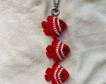 May include: A red and white crocheted fish keychain with a silver metal clasp. The fish are connected in a line, with the top fish attached to the clasp.