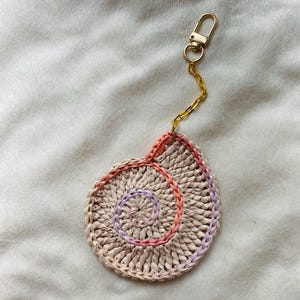 May include: A handmade, beige seashell-shaped keychain. The spiral design is accented with pink and purple thread. The keychain has a gold-colored clasp and chain.