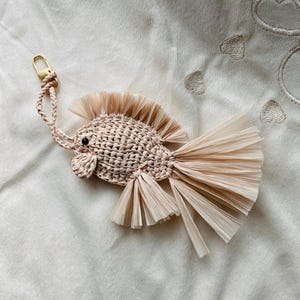 May include: A beige macrame fish keychain with a gold-colored clasp. The fish has a textured body, a fringed tail, and a fin-like detail on its back. The keychain is lying on a soft, off-white surface.