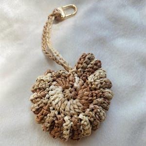 Pode incluir: Um chaveiro artesanal com um pingente em forma de flor tecida em tons de marrom e bege. O pingente é preso a um cordão trançado e a um fecho dourado. O fundo é um tecido branco e macio. O diâmetro é de aproximadamente 6 cm.
