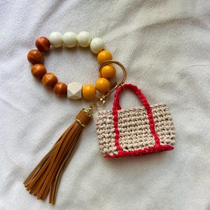 May include: A beaded keychain bracelet with a tan leather tassel and a small, crocheted tote bag charm with red trim. The bracelet features wooden beads in shades of brown, white, and yellow, connected by a gold-tone ring.