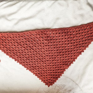 Op de afbeelding: Een roestkleurige gehaakte driehoekige sjaal met twee lange kwastjes. De sjaal is gemaakt met een eenvoudig steekpatroon en heeft een delicate, kantlook.