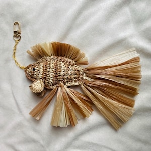 May include: A woven fish keyring in shades of brown and beige. The fish has a detailed woven body, a fringed tail, and a fin. A gold-coloured chain and clasp are attached to the fish's head.