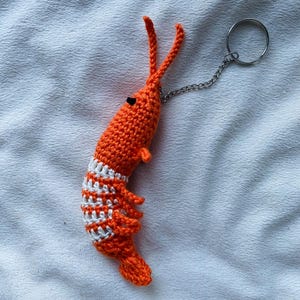 May include: A handmade orange crochet shrimp keychain with white stripes and a silver chain and ring. The shrimp has two long antennae and small black eyes. The keychain is a unique, whimsical accessory.