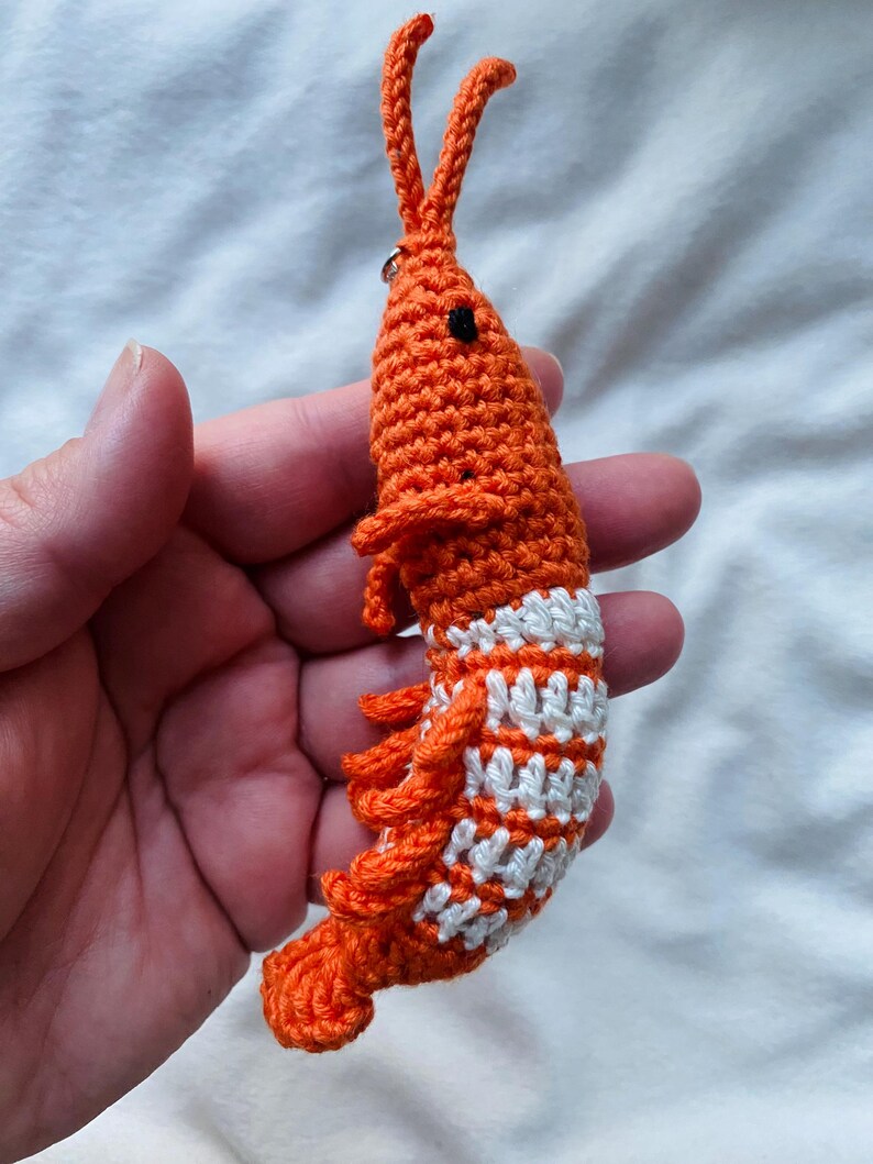 May include: A handmade orange and white crochet shrimp keychain. The shrimp has a detailed design with small black eyes, white stripes, and orange fringe. The keychain is held in a hand against a white background.