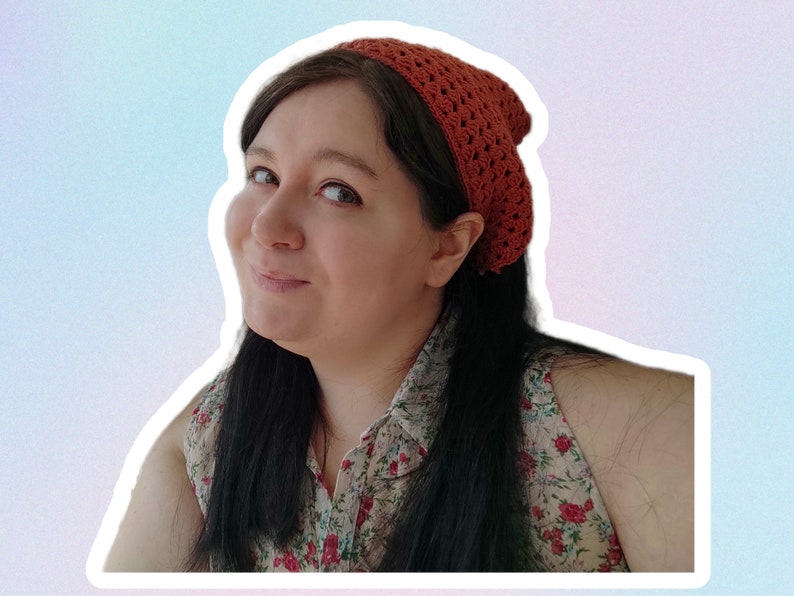Op de afbeelding: Een vrouw met lang donker haar draagt een gehaakt hoofdband in een verbrand oranje kleur. Ze draagt een mouwloos blouse met een bloemenpatroon.