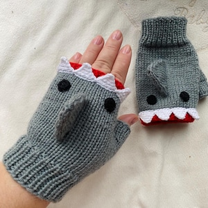May include: Grey knitted shark mittens with white teeth, black eyes, and a red interior. One mitten is on a hand, the other is next to it. The mittens have a fingerless design.
