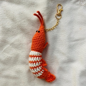 May include: A handmade orange crochet shrimp keychain with a white and orange striped midsection. The shrimp has black eyes and two orange antennae. It is attached to a gold-colored metal clasp and chain. The keychain is a whimsical accessory.
