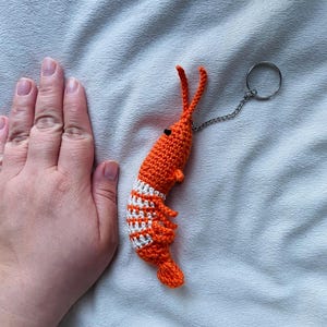 May include: A handmade orange and white crochet shrimp keychain. The shrimp has a detailed design with white stripes and a small chain attached to a keyring. The keychain is a unique accessory.