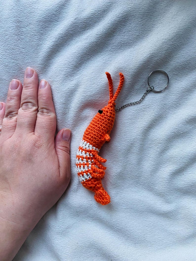 May include: A handmade orange and white crochet shrimp keychain. The shrimp has a detailed design with white stripes and a small chain attached to a keyring. The keychain is a unique accessory.