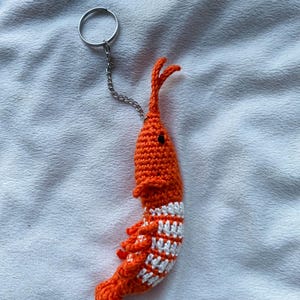 May include: A handmade orange crochet shrimp keychain with white stripes on its body. The shrimp has small black eyes and a silver keyring attached with a chain. The keychain is a unique accessory.