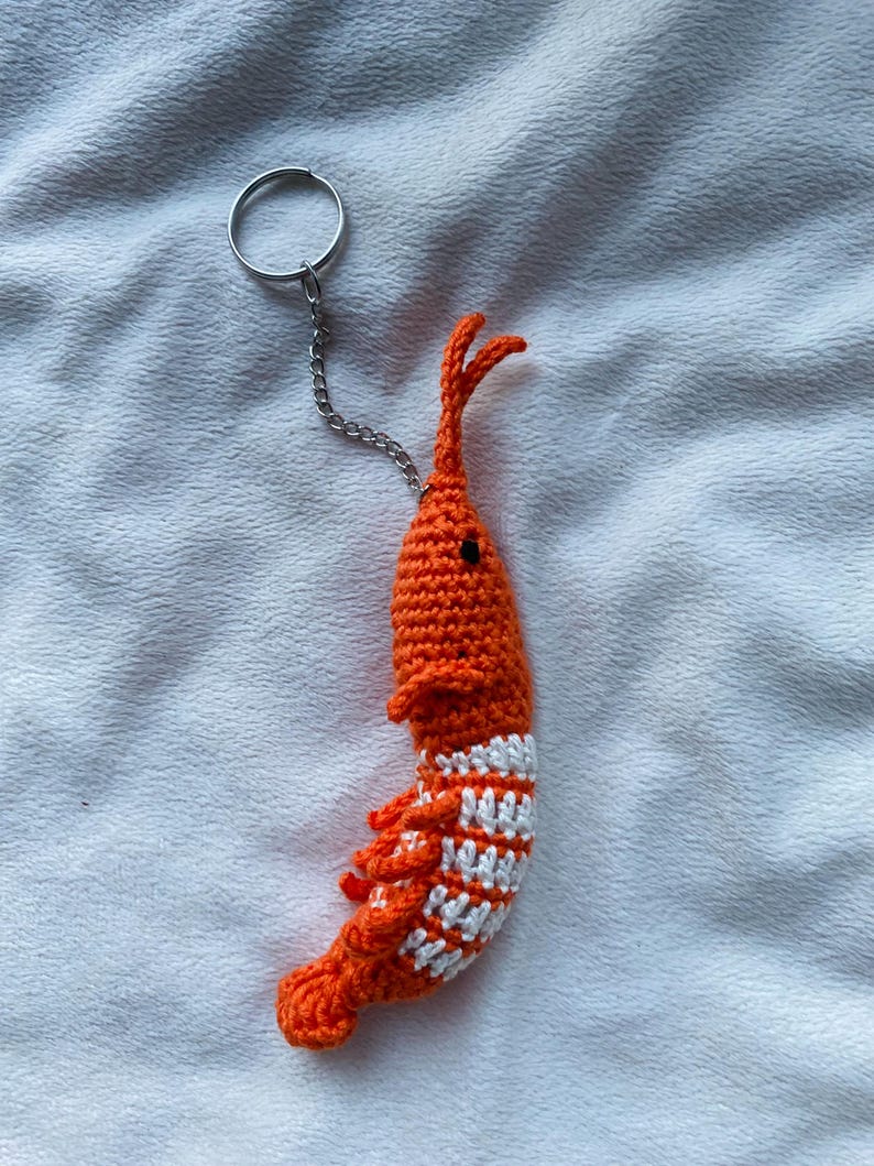May include: A handmade orange crochet shrimp keychain with white stripes on its body. The shrimp has small black eyes and a silver keyring attached with a chain. The keychain is a unique accessory.