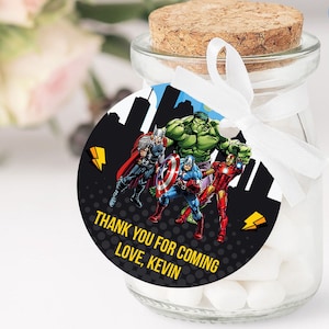 Puede incluir: Un pequeño tarro de cristal lleno de caramelos blancos, coronado con un corcho y una etiqueta redonda. La etiqueta presenta ilustraciones de superhéroes, una silueta de ciudad y el texto "THANK YOU FOR COMING LOVE, KEVIN". Una cinta blanca está atada alrededor del tarro.