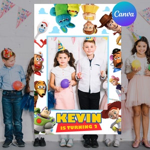 May include: A photo booth frame featuring characters from the Toy Story movie, with the text "KEVIN IS TURNING 2". The frame is decorated with images of Woody, Buzz Lightyear, and other characters. Children are posing with balloons in front of the frame.