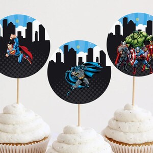 Puede incluir: Tres cupcakes con adornos temáticos de superhéroes. Cada adorno presenta un superhéroe diferente: Superman, Batman y un grupo de Vengadores. Los adornos tienen una silueta de paisaje urbano negro sobre un fondo de cielo azul.