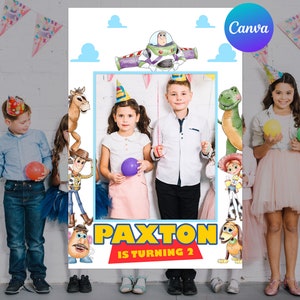 May include: A custom photo booth prop featuring Toy Story characters and the text "PAXTON IS TURNING 2". The design includes Buzz Lightyear, Woody, and other characters, with a blue and white cloud background. Children are holding balloons.