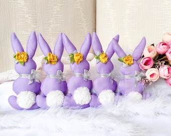 May include: Five purple fabric Easter bunnies with yellow flower accents and white pom-poms. The bunnies are lined up on a white fluffy surface.