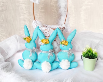 May include: Three turquoise bunny figurines with white fluffy tails, yellow flower accents, and silver ribbon bows. A white lace-trimmed basket with a brown handle and a white lace bow are in the background. A small green bucket with faux greenery is on the right.