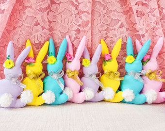 May include: Nine pastel colored plush bunny rabbits with embroidered flower accents. The bunnies are arranged in a row, with the pink bunny in the center.