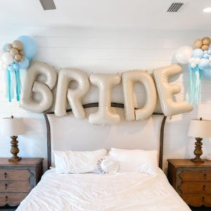 Puede incluir: Una cama blanca con una cabecera blanca y un edredón blanco. La cabecera tiene una gran pancarta de globos beige que deletrea "BRIDE". Hay globos azules y beige a cada lado de la pancarta.