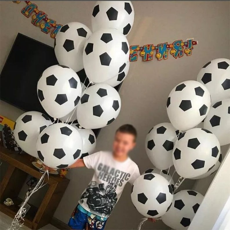 Może przedstawiać: Grupa biało-czarnych balon&oacute;w w kształcie piłki nożnej. Balony są związane ze sobą i trzymane przez dziecko w czarno-białej koszulce z napisem "Galactic Heroes".