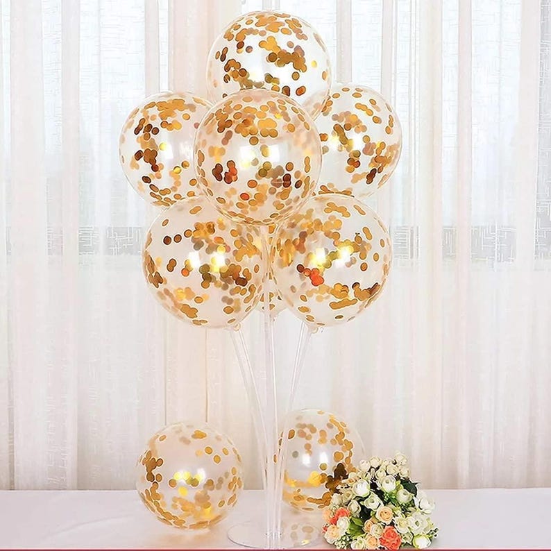 May include: A cluster of clear balloons with gold confetti inside, arranged on a clear stand. The balloons are tied together with clear string.