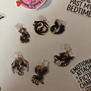 May include: A selection of black and gold metal charms depicting dragons, phoenixes, and ravens. Each charm has a small clear plastic clip. Decorative stickers with text are also visible, including "I READ PAST MY BEDTIME".