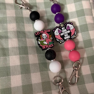 May include: Two beaded keychains with silver clasps. One has black, white beads, and a charm that says "Let Me Read In Peace". The other has purple, pink beads, and a charm that says "My Book Boyfriend Has Wings".