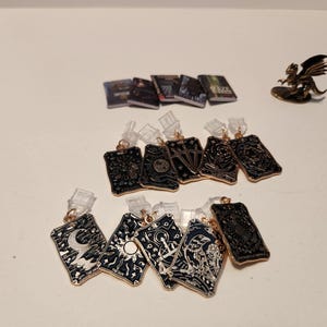 May include: A collection of miniature items, including small books, tarot card pendants, and a dragon figurine. The pendants have intricate black and gold designs, with various symbols. The dragon figurine is gold-coloured.