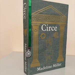 Op de afbeelding: Een hardcover boek getiteld "Circe" van Madeline Miller. Het boek heeft een groene kaft met gouden architecturale ontwerpen en tekst. Een groen lint-bladwijzer met een zilveren bedel is ingevoegd. Het boek staat op een wit oppervlak.