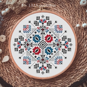 May include: A white embroidery hoop featuring a colourful geometric pattern in red, blue, and black. The pattern is a traditional folk design with a central square and four corner motifs.