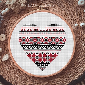 May include: A red, black, and white cross-stitch pattern of a heart with a traditional folk design. The heart is framed in a wooden embroidery hoop.
