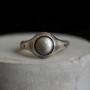 May include: A silver ring featuring a prominent, circular central element. The ring has an unusual, organic form with a textured finish. The band narrows towards the rear, and the overall design is minimalist and contemporary.
