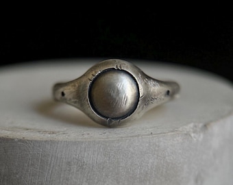May include: A silver ring with a large, circular center stone. The ring has a unique, organic shape with a textured surface. The band tapers towards the back, and the overall design is minimalist and modern.