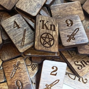 Puede incluir: Primer plano de un montón de fichas de madera con varios números, símbolos y letras, incluyendo "Kn" y un pentagrama. Las fichas están teñidas de un color marrón oscuro y tienen un aspecto rústico.