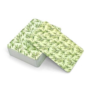 Op de afbeelding: Een kaartspel met een bamboebladpatroon. De kaarten, groen en wit, zitten in een zilveren metalen doos. De kaarten zijn iets uitgespreid en tonen het ontwerp op de achterkant.