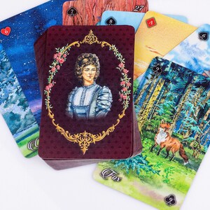 Op de afbeelding: Een tarotkaartspel met diverse aquarel illustraties. De centrale kaart toont een portret in een decoratief kader. Andere kaarten tonen sc&egrave;nes uit de natuur, waaronder een vos, en hemellandschappen. De kaarten zijn gerangschikt op een wit oppervlak.