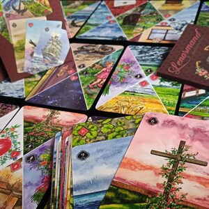 Op de afbeelding: Een Lenormand solitaire kaartspel met een bruine doos en kaarten met aquarel illustraties. De kaarten tonen verschillende afbeeldingen, waaronder een hart, een kruis en landschappen. De doos heeft de naam "Lenormand" erop.