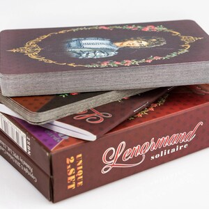 Op de afbeelding: Een stapel Lenormand solitaire kaartspellen. Het bovenste kaartspel heeft een portret in een decoratief gouden frame. De dozen zijn bordeauxrood met witte tekst en de woorden "Lenormand solitaire" zijn zichtbaar.
