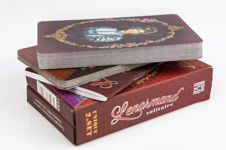 Op de afbeelding: Een stapel Lenormand solitaire kaartspellen. Het bovenste kaartspel heeft een portret in een decoratief gouden frame. De dozen zijn bordeauxrood met witte tekst en de woorden "Lenormand solitaire" zijn zichtbaar.